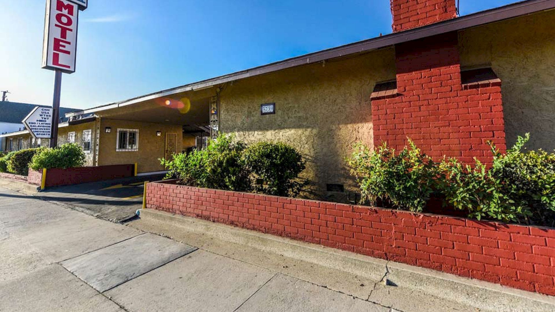 The image shows the exterior of a single-story motel with a "Motel" sign, beige walls, red brick chimney, and landscaped bushes in front.